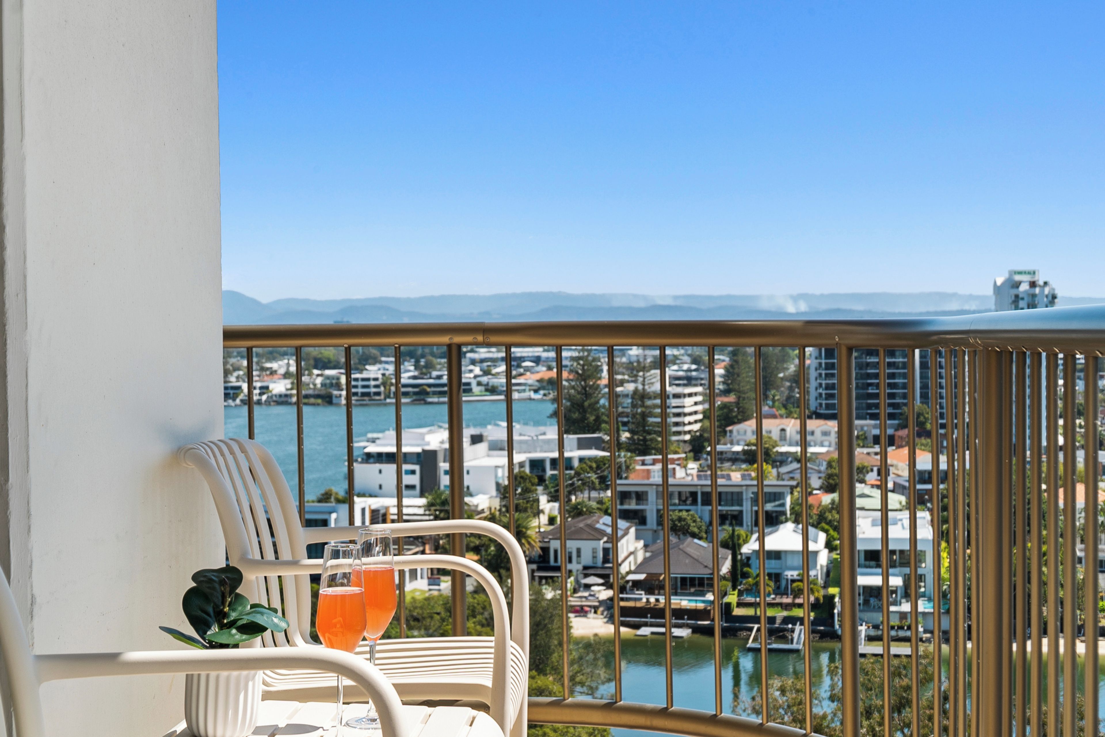 A balcony with a view of a marina and buildings, a white chair, and a glass table with a drink.