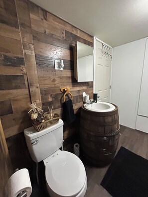 A bathroom with a wooden wall, a toilet, a sink with a wooden barrel, and a mirror. - Cozy cottage cabin with lake and beach access (Albion)