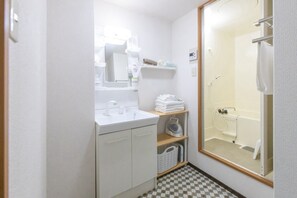 Een kleine, schone badkamer met een witte wastafel, spiegel en douche. Er staan planken met handdoeken en toiletartikelen.