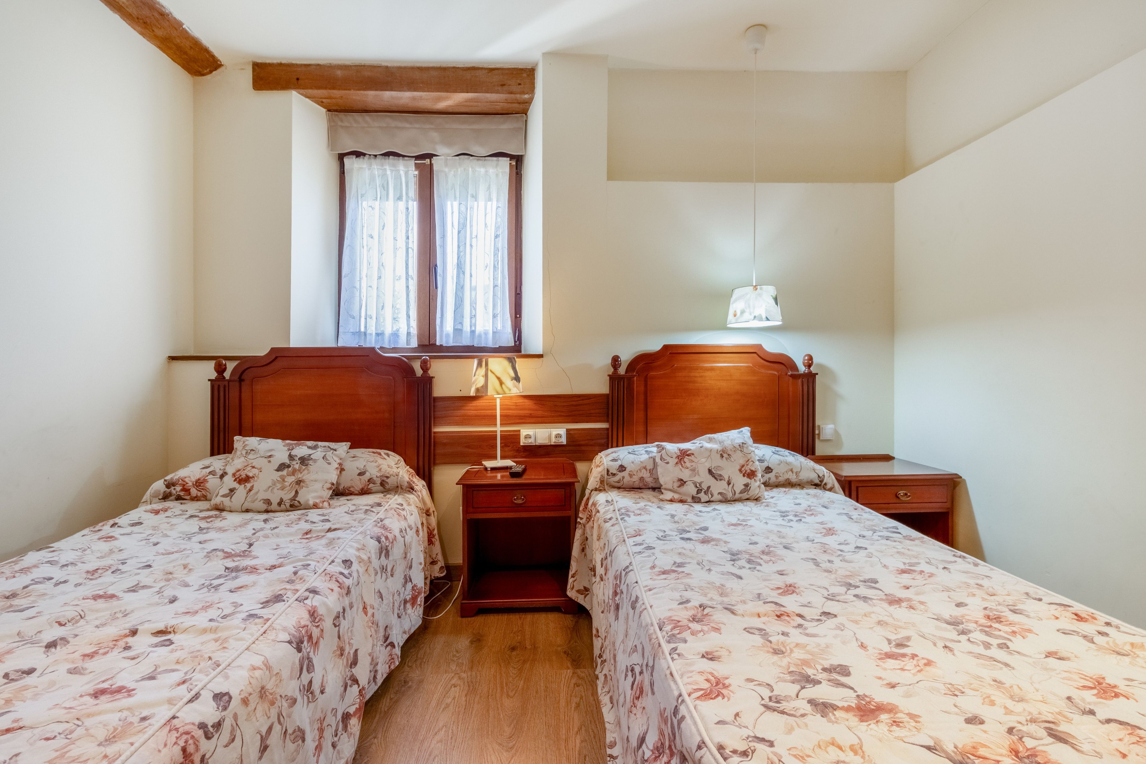 Un dormitorio con dos camas de madera, una mesita de noche con lámpara y una ventana con cortinas.