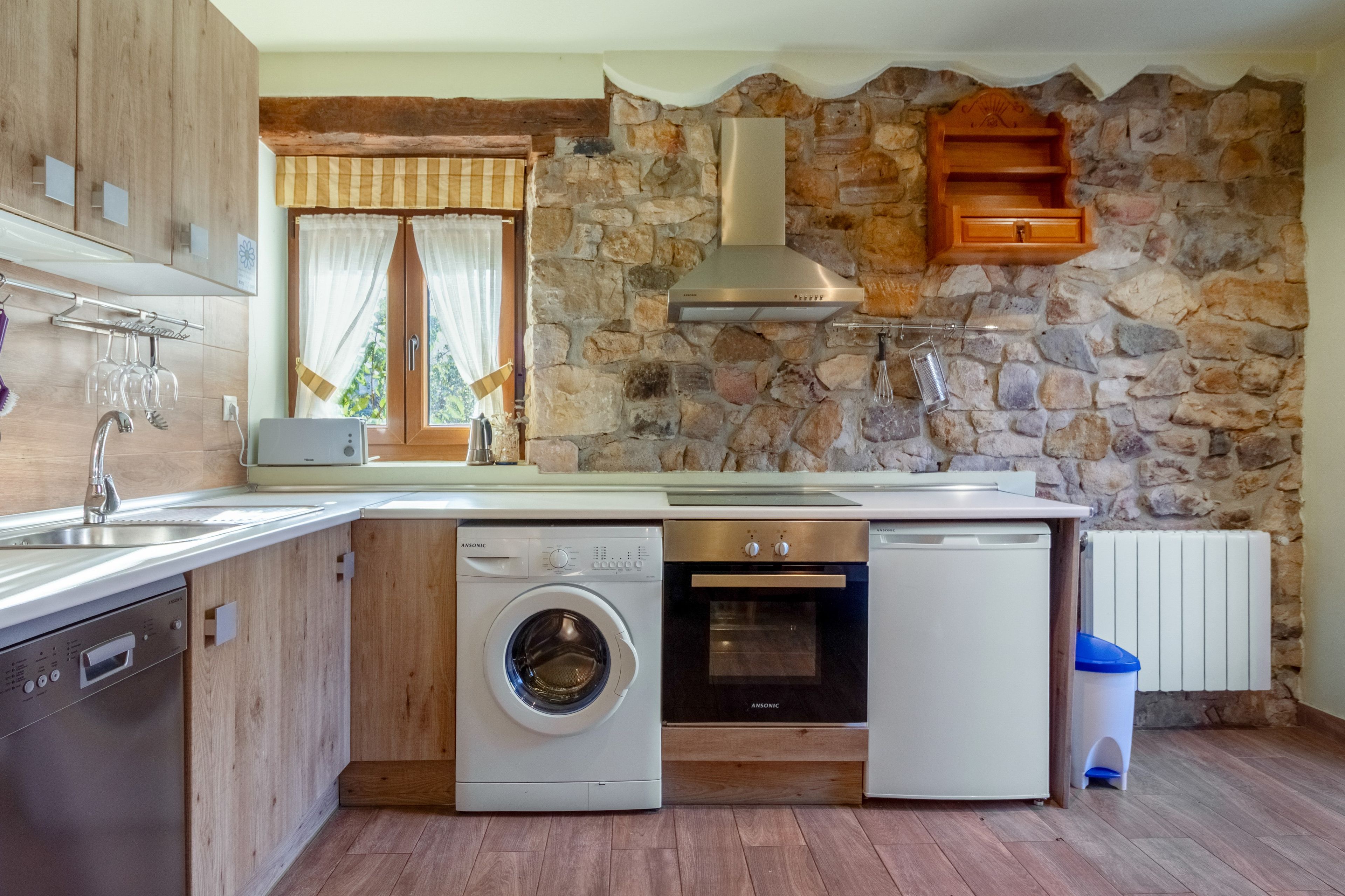 Cocina compacta con pared de piedra, armarios de madera, horno integrado, lavavajillas y lavadora.