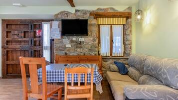Una sala de estar acogedora con una pared de piedra, un televisor sobre la chimenea, una mesa de comedor de madera con sillas y un sofá.