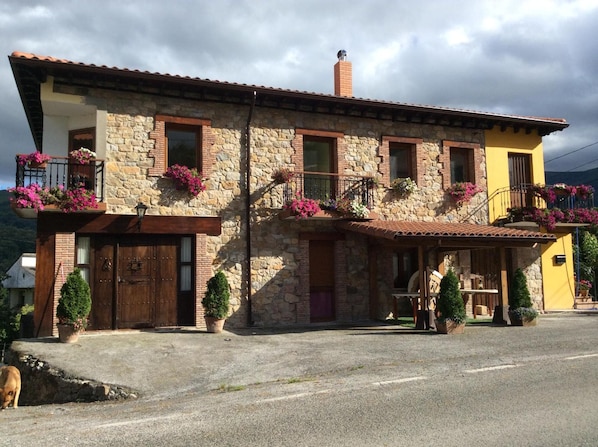Edifício de pedra de dois andares com varandas decoradas com flores, uma porta de madeira e uma pequena área de estar ao ar livre.