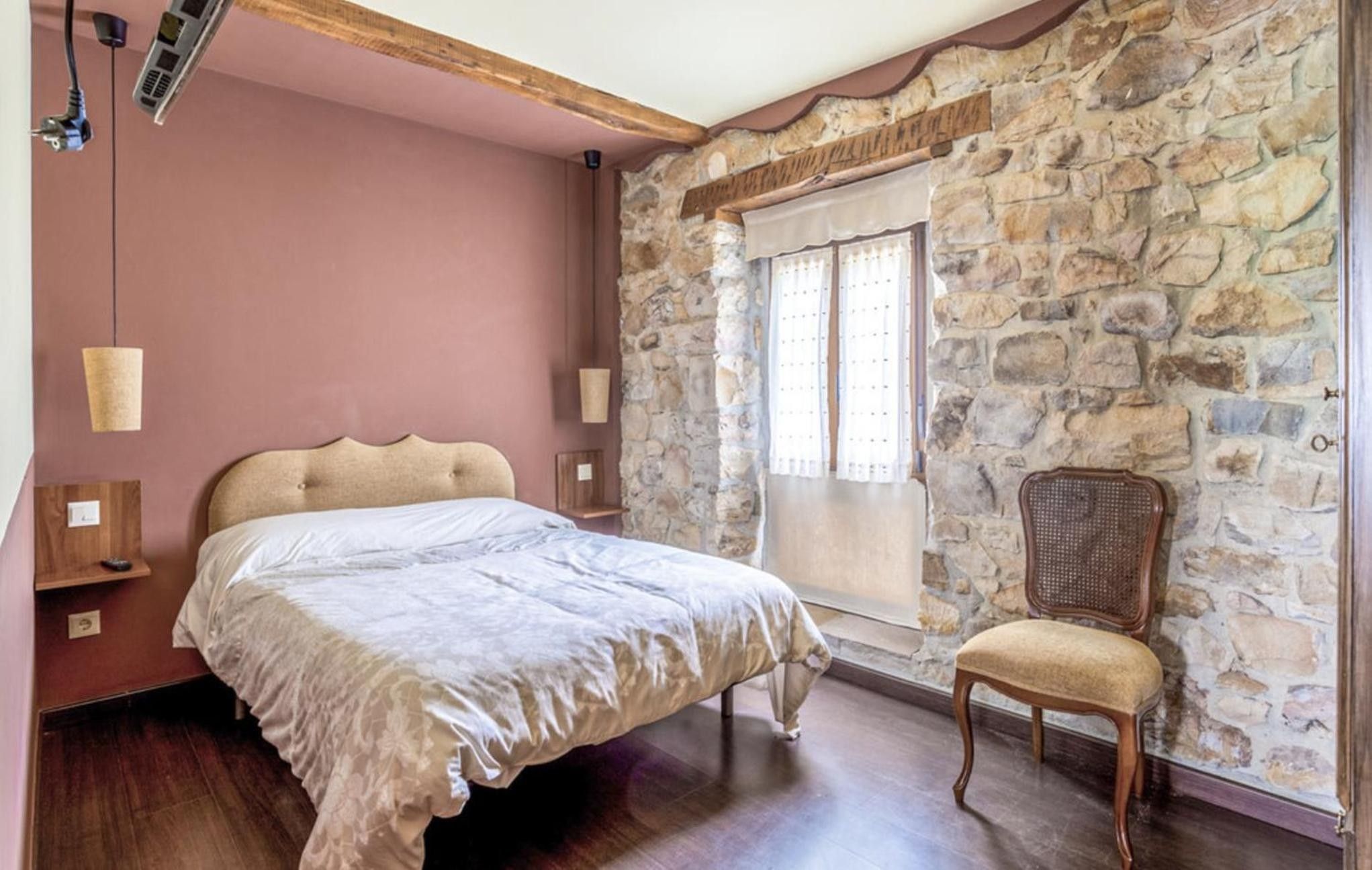 A bedroom with a stone wall, a bed, a chair, and a window with curtains.