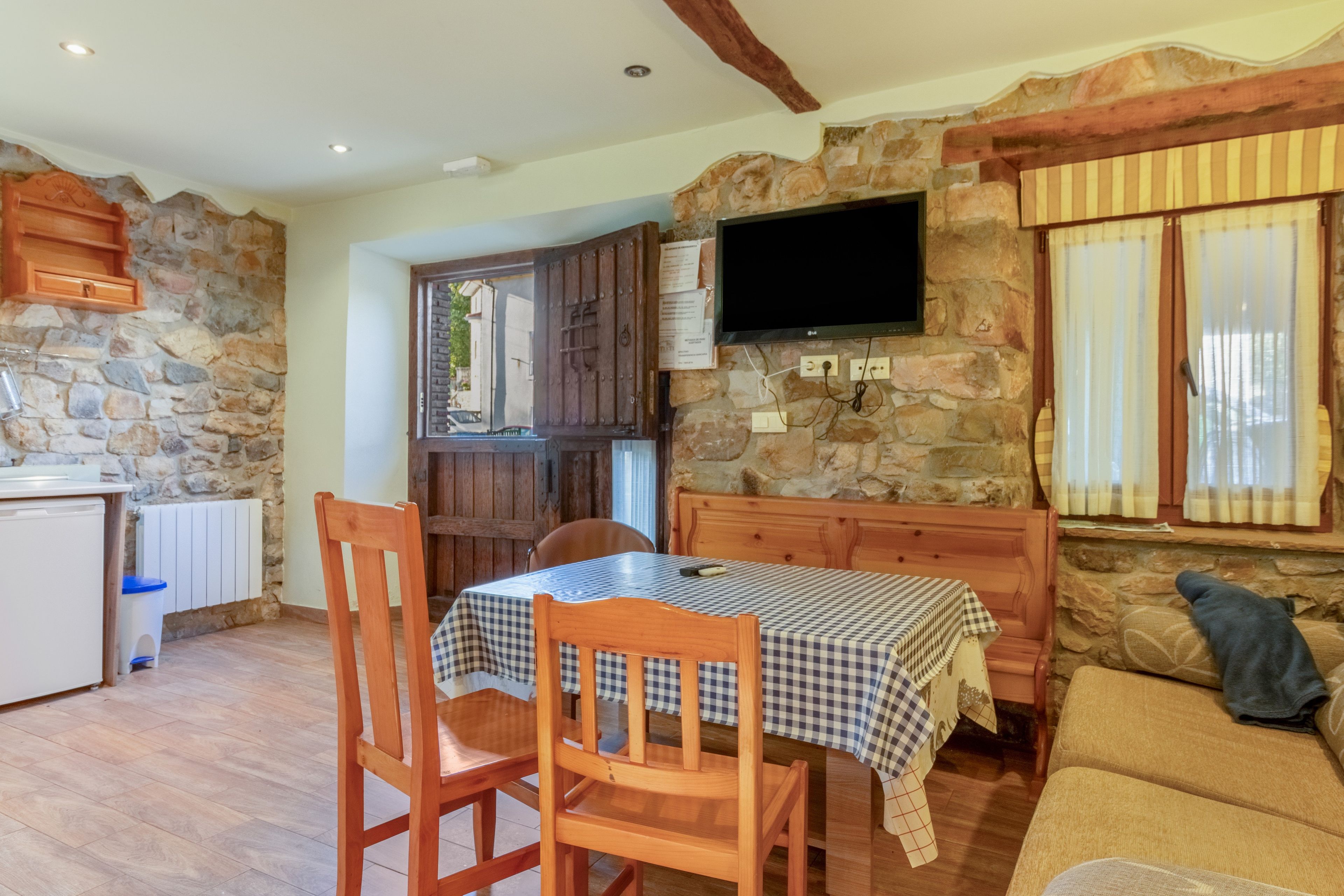 A cozy room with a stone wall, a flat-screen TV mounted on the wall, a wooden table with a checkered tablecloth, and a sofa with a cushion.