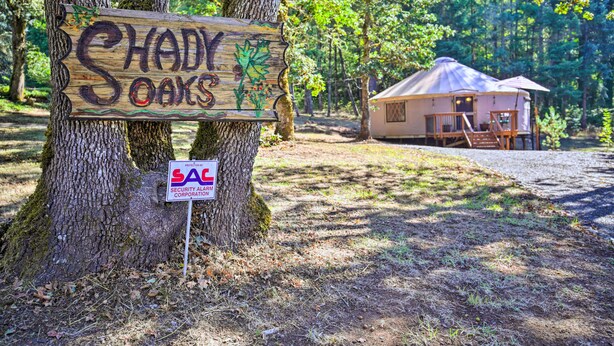 Un letrero de madera con la inscripción 'SHADY OAKS' y un letrero de 'SAC SECURITY ALARM CORPORATION', con una estructura tipo yurt en el fondo.