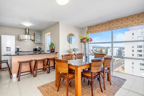A modern kitchen with a dining area, large windows, and a view of the city. - The Waves 704 - Two Bedroom Apartment, Sleeps 4 (Cape Town)