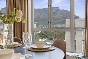 Un coin repas avec une table dressée pour deux personnes, un vase rempli de fleurs et une vue sur une montagne et une ville.