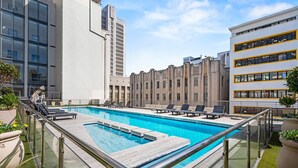 Pool på taget med liggestole og glasgelænder, omgivet af moderne bygninger.