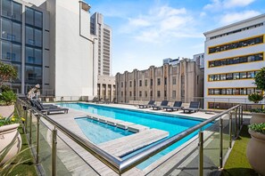 Une piscine sur le toit, dotée de chaises longues et d’une rambarde en verre, et entourée d’immeubles modernes.
