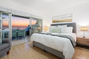 Ein modernes Hotelzimmer mit einem großen Bett, einem Schreibtisch mit Stuhl, einer Couch und Blick auf den Sonnenuntergang.