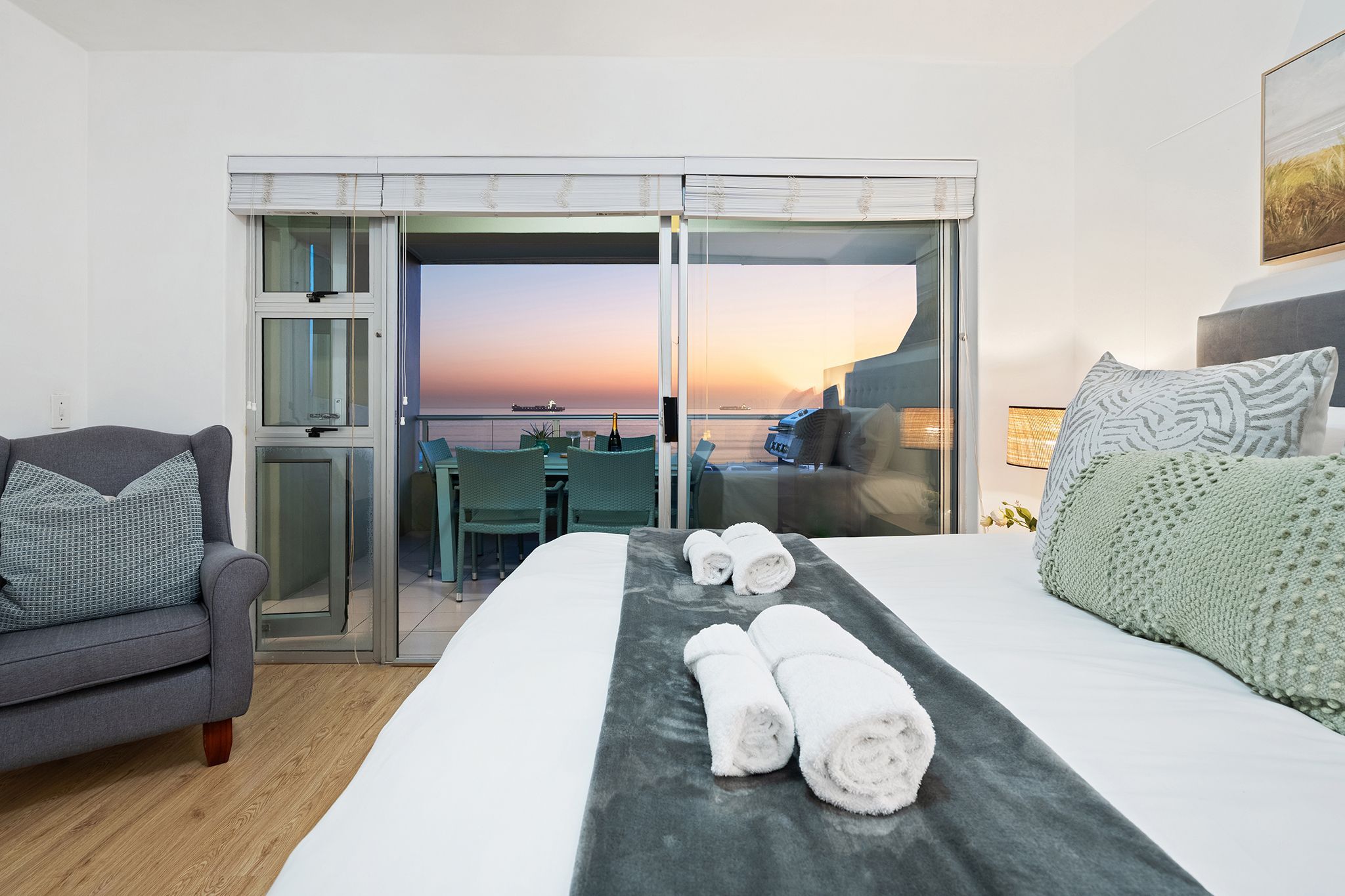 A hotel room with a bed, a sofa, a dining area with a table and chairs, and a view of the sea at sunset.