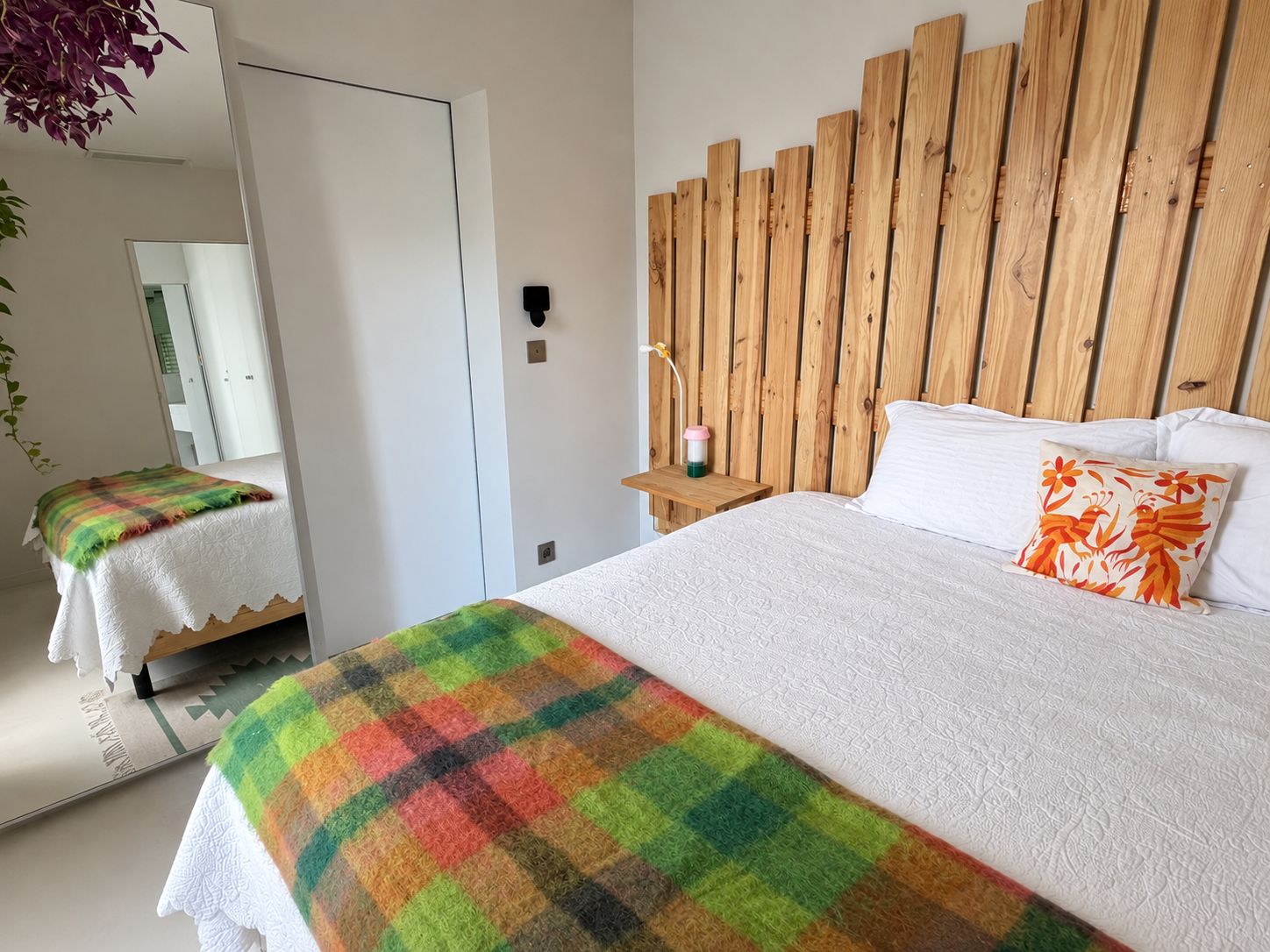 A bedroom with a wooden headboard, a bed with white bedding, a colorful blanket, and a nightstand with a lamp.
