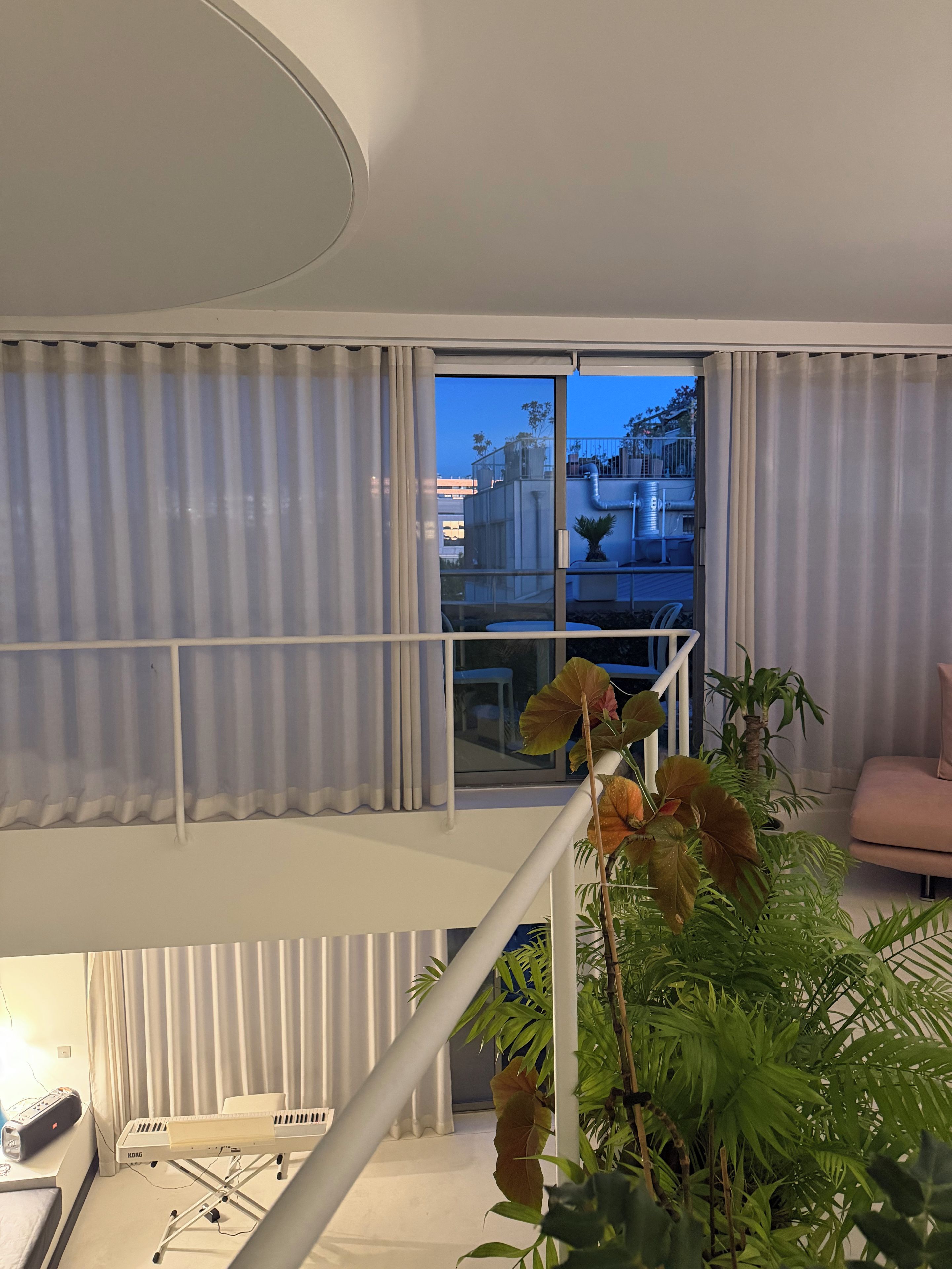 A modern living room with a view of the cityscape through large windows, a white staircase, and a variety of indoor plants.