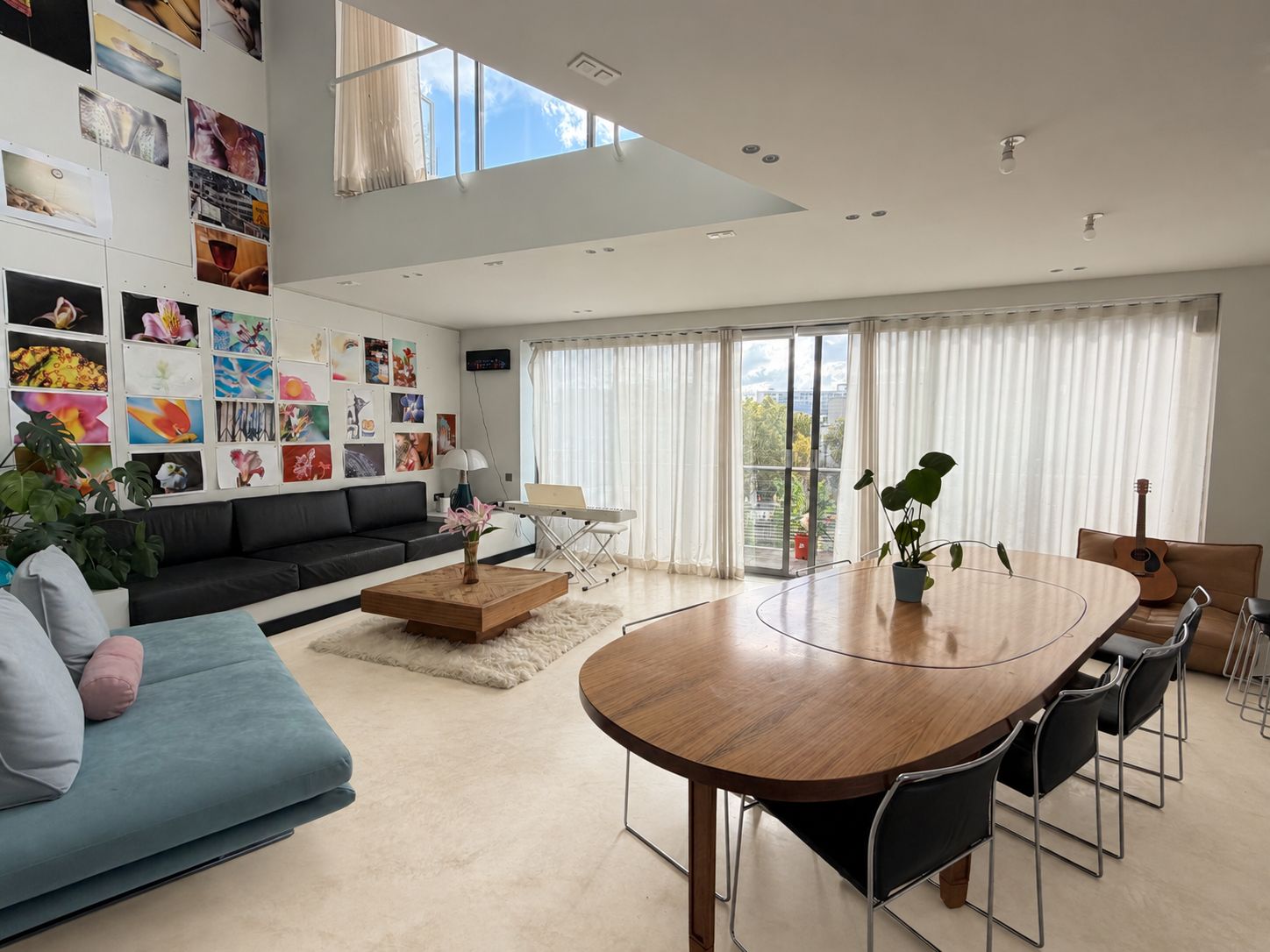 Een moderne woonkamer met een bank, salontafel en eettafel met stoelen. Er is een groot raam met gordijnen, een muur met kunstwerken en een gitaar in de hoek.
