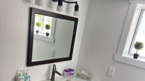 Un baño con tocador blanco, un espejo, una lámpara de pared negra y una ventana con una planta en maceta.