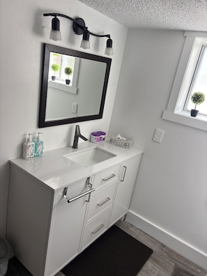 Een badkamer met een wit wastafelmeubel, een spiegel, een zwart wandlampje en een raam met een bloempot.