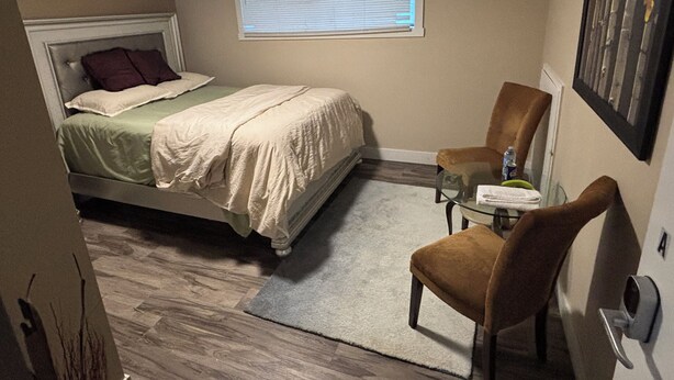 Un dormitorio con cama, una silla, una mesita, un ventanal y una puerta.