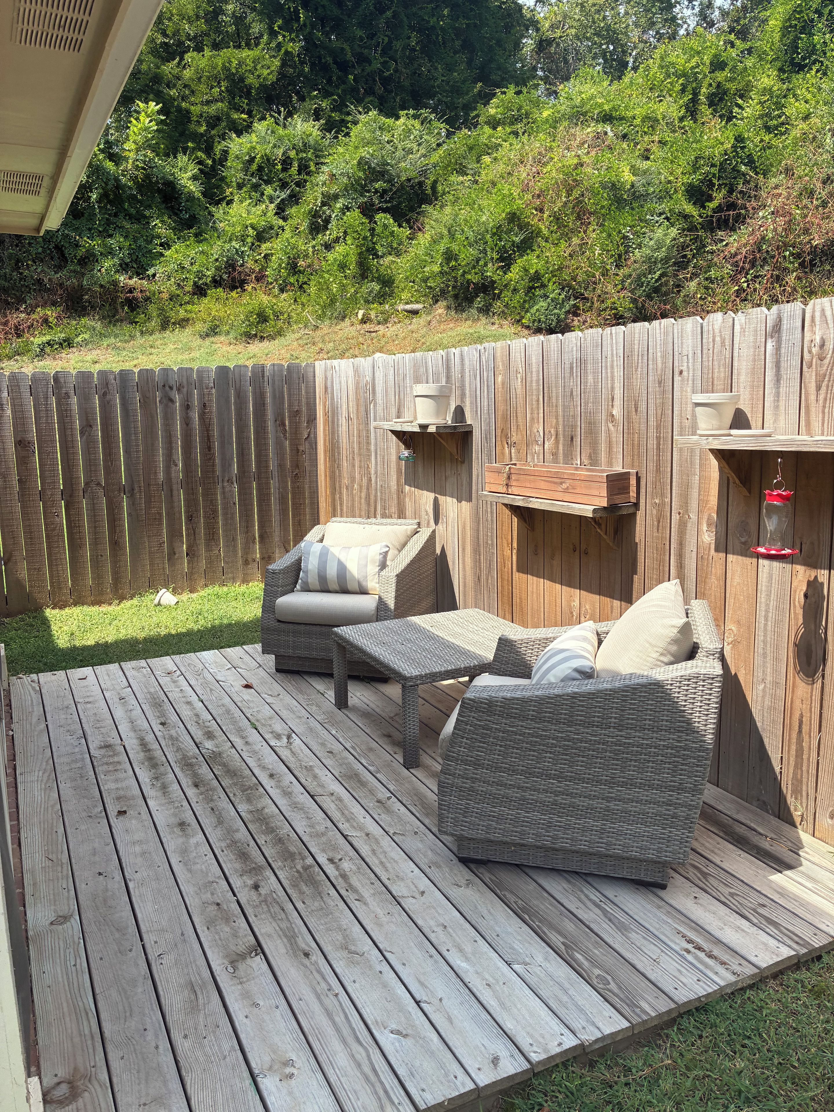 Una terraza de madera con mobiliario de mimbre, incluyendo un sofá y una mesa, y una valla de madera al fondo.