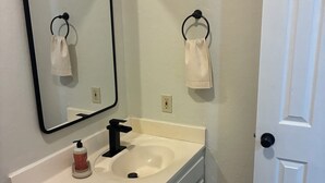 Uma casa de banho com lavatório branco, espelho, suporte de toalha, rolo de papel higiénico e porta.