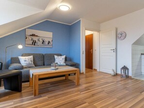 Un salon avec un canapé, une table basse et une lampe. Il y a une porte qui donne sur une autre pièce et une horloge au mur.