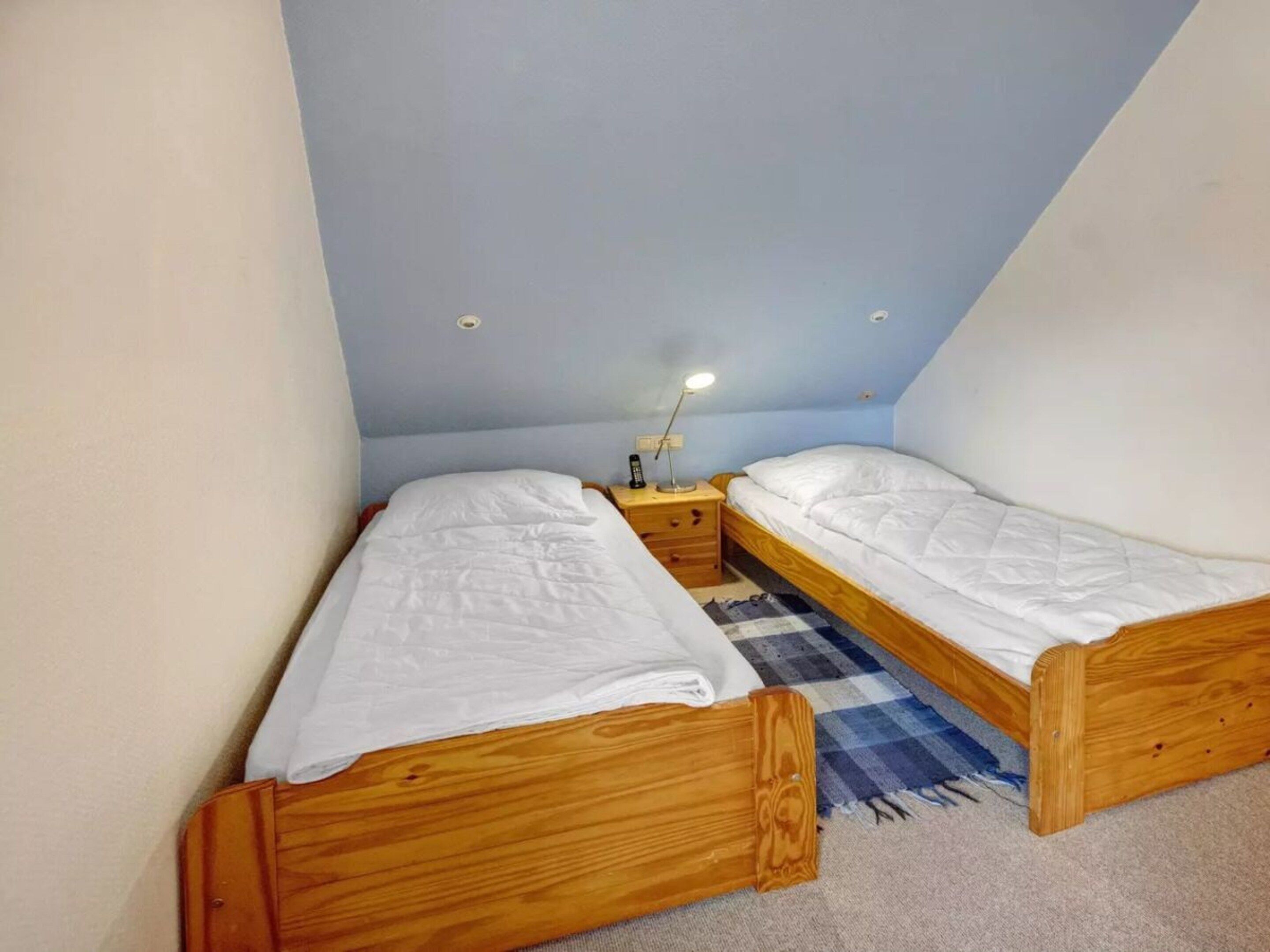 A small, single-room accommodation with two wooden beds, a nightstand with a lamp, and a checkered rug on the floor.