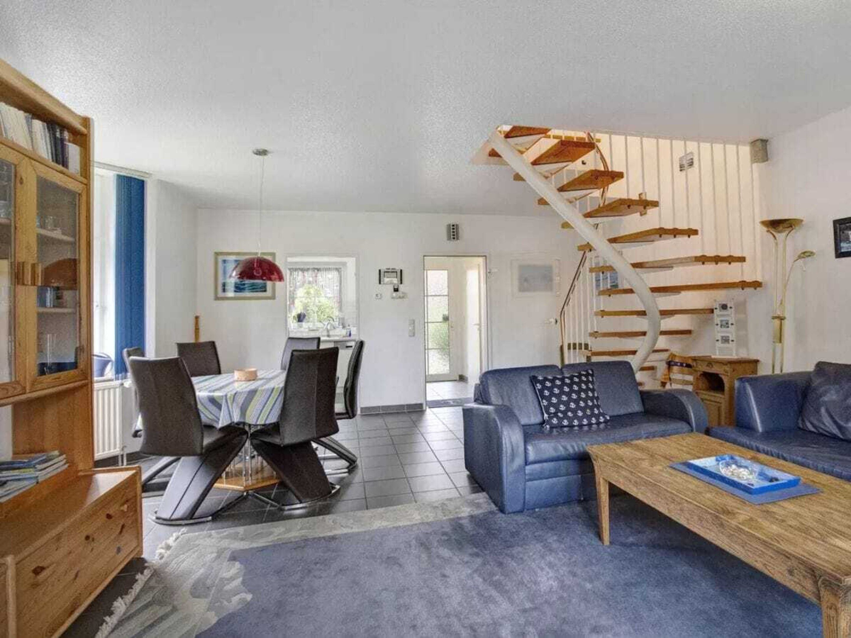 A living room with a blue sofa, a coffee table, a dining area with chairs, a staircase, and a bookshelf.