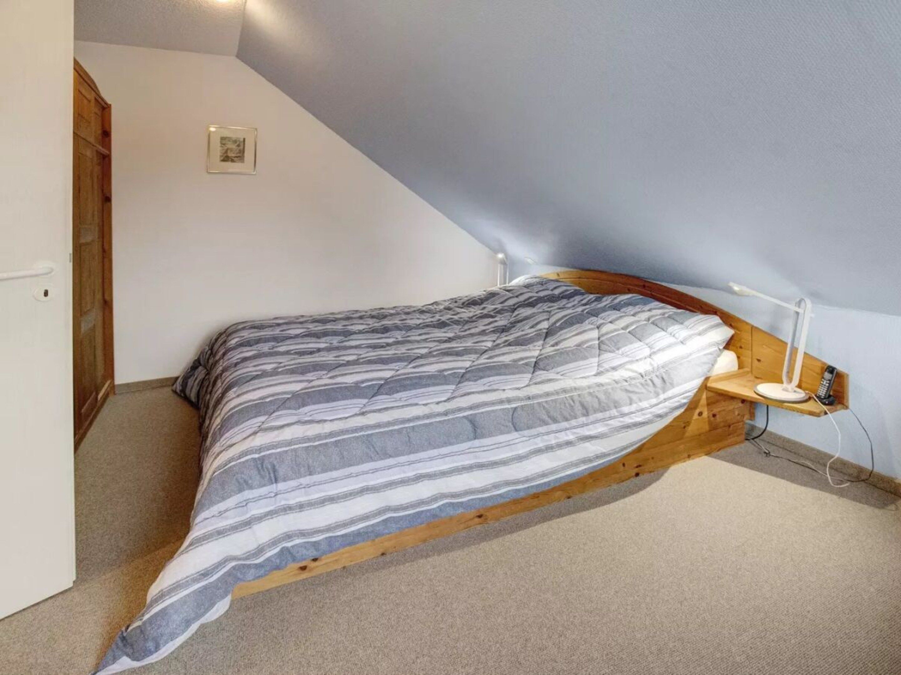 Ett sovrum med ett sluttande tak, en säng med ett randigt överkast, en sänglampa och en liten hylla med en telefon på.