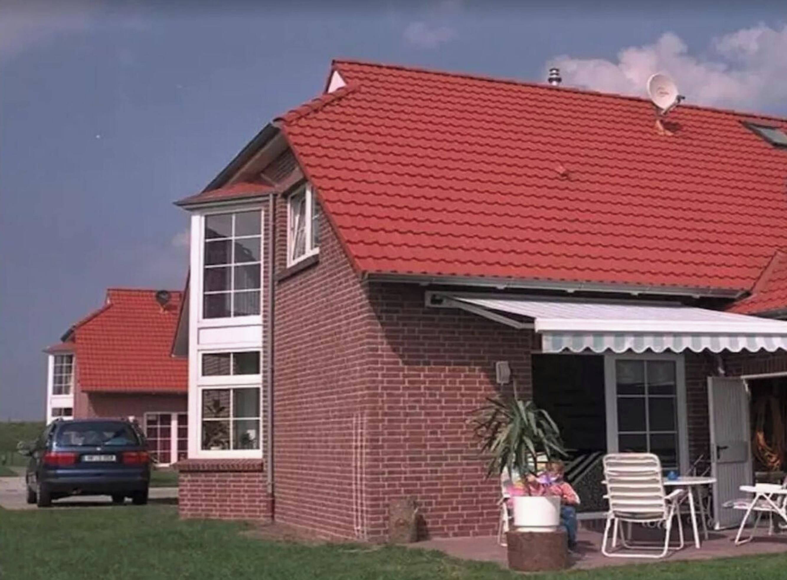 A two-story brick house with a red tiled roof, a covered patio area with outdoor furniture, and a car parked in the driveway.
