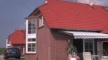 A two-story brick house with a red tiled roof, a covered patio area with outdoor furniture, and a car parked in the driveway.