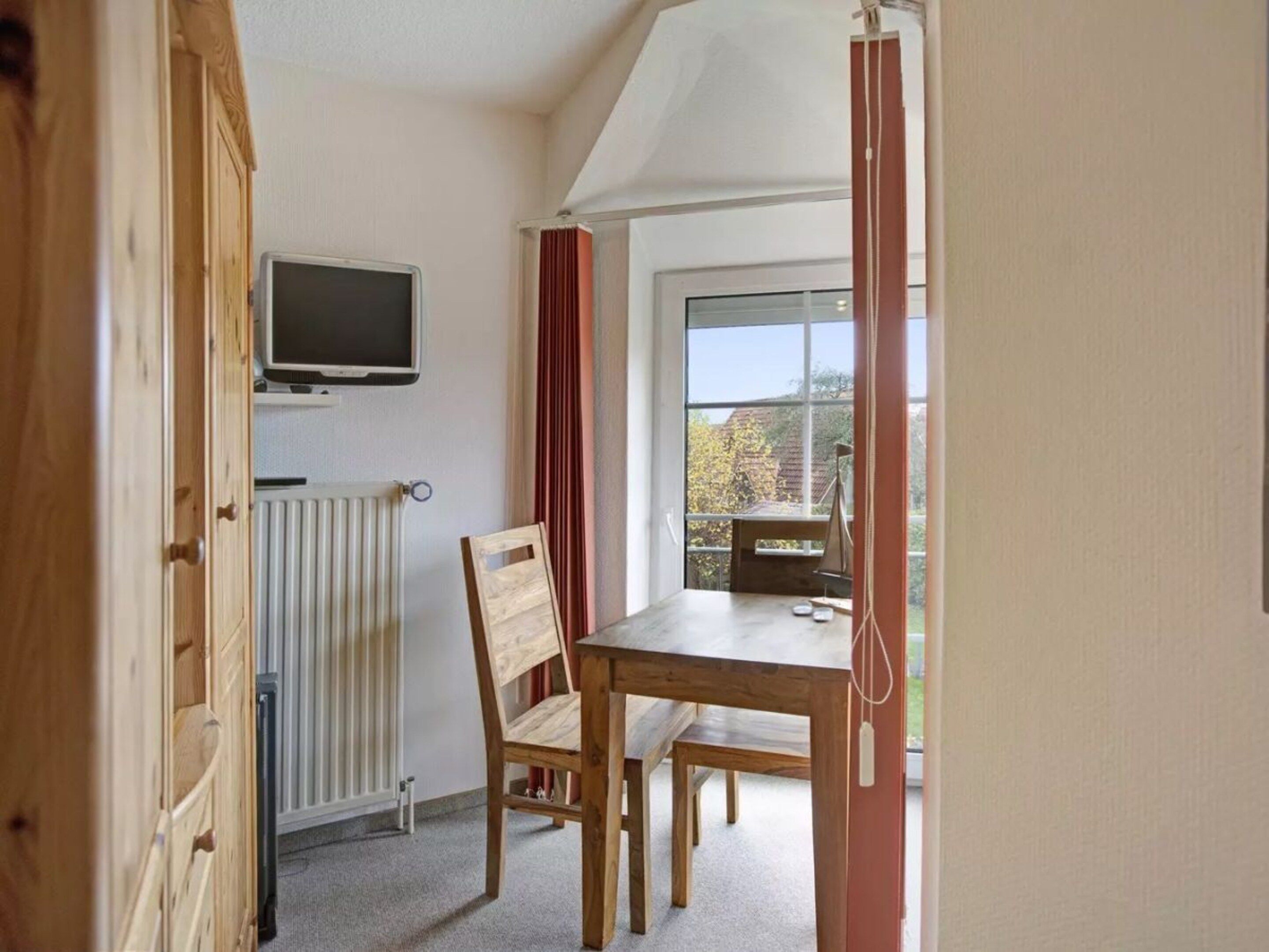 A room with a wooden table and chair, a television mounted on the wall, and a window with a view of trees.
