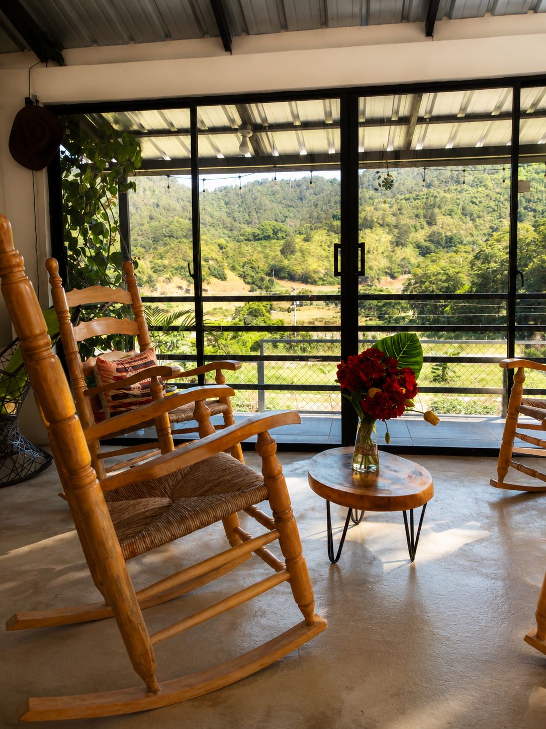 Una habitación con ventanas amplias, mecedoras de madera, una mesita redonda con un florero de flores rojas y vistas a un paisaje verde.