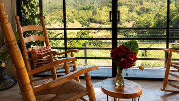 Una habitación con ventanas amplias, mecedoras de madera, una mesita redonda con un florero de flores rojas y vistas a un paisaje verde.