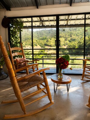 A room with large windows, wooden rocking chairs, a small round table with a vase of red flowers, and a view of a green landscape.