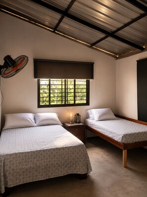 Un dormitorio con dos camas, un ventilador de pie, una mesita de noche con una lámpara y una ventana con vista a follaje.