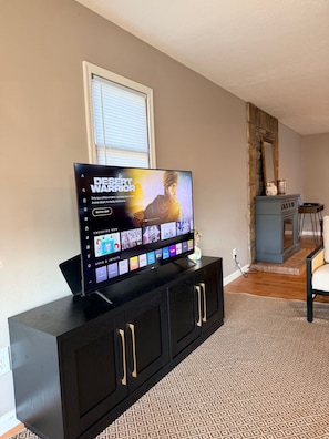 A flat-screen TV on a black entertainment unit with a desert warrior movie playing, a window with blinds, and a stone fireplace. - Newly Renovated 2BR Retreat – Minutes from CVG & Cincinnati (Petersburg)