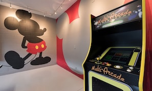 Ein Mehrfach-Arcade-Spielgerät mit farbigem Bildschirm und Bedienfeld, vor einer Wand mit einem großen Wandgemälde von Micky Maus.
