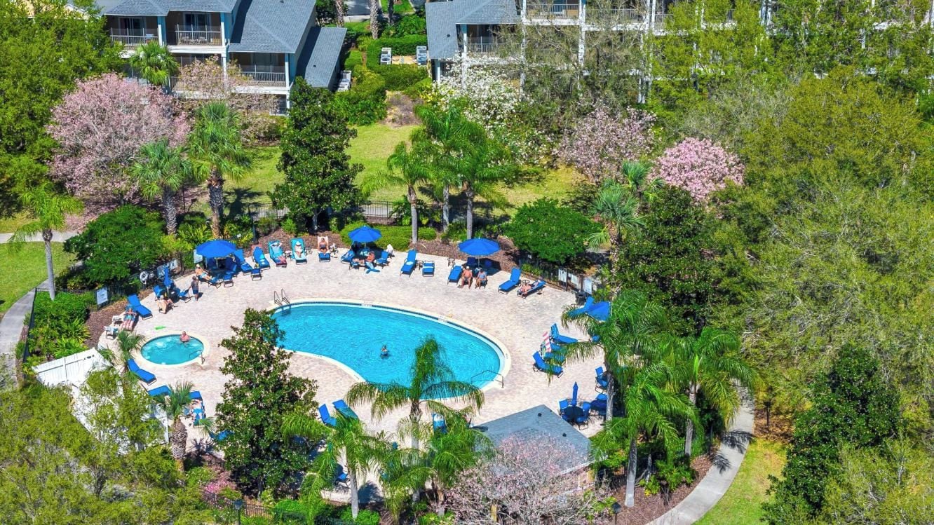 An aerial view of a resort with a central swimming pool surrounded by lounge chairs and umbrellas, with lush greenery and buildings in the background.
