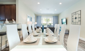 A dining area with a long table set for multiple people, a kitchen area with cabinets, and a living room with a television. - 6BR Encore Villa Themed Rooms & Private Pool (Kissimmee)