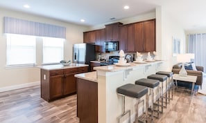 A modern kitchen with dark wood cabinets, a central island with bar stools, a refrigerator, and a microwave. - 6BR Encore Villa Themed Rooms & Private Pool (Kissimmee)