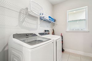 A laundry area with a front-loading washer and dryer, a metal shelf with folded clothes, a window with blinds, and a vacuum cleaner. - 5BR Bear's Den Estate Private Pool & Game Room (Kissimmee)