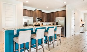 A modern kitchen with dark wood cabinets, stainless steel appliances, a blue accent wall, and three bar stools. - 9BR Encore Villa Custom Design & Game Room (Kissimmee)