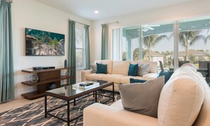 A modern living room with a beige sofa, a glass coffee table, and a TV mounted on the wall. - 5BR Encore Villa With Billiards Loft & Pool (Kissimmee)