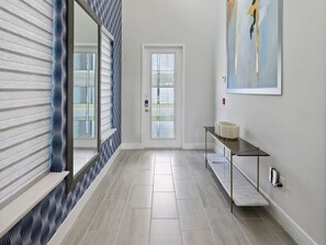 A hallway with a patterned wall, a door with frosted glass panels, a console table with a vase, and a painting on the wall. - 7BR Luxe Mansion Private Pool & Golf Views (Kissimmee)