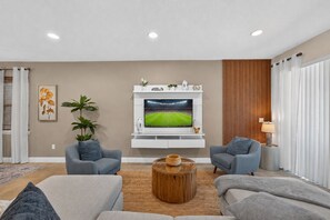 A modern living room with a flat-screen TV mounted on a white wall, two grey sofas, a wooden coffee table, and a large window with vertical blinds. - 6BR Encore w/ Under-the-sea Kids' Room & Pool (Kissimmee)