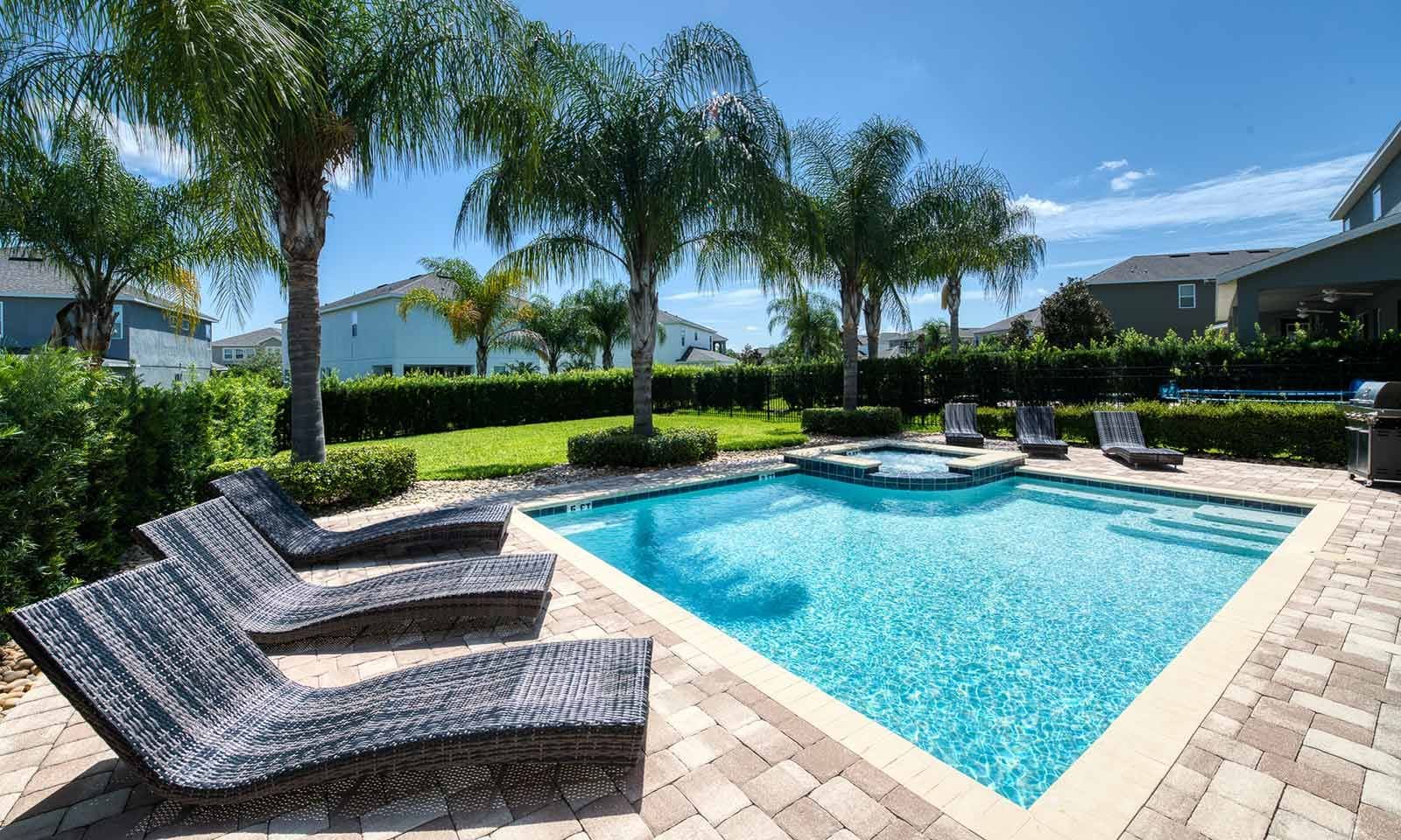 Una piscina rectangular con una bañera integrada, rodeada por un patio de ladrillo y palmeras.