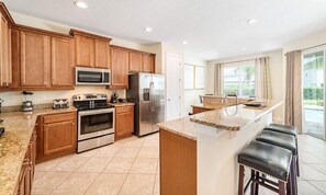 A modern kitchen with wooden cabinets, stainless steel appliances, a granite countertop, and a bar area with stools. - 6BR Encore Villa Pool, Spa & Game Room (Kissimmee)