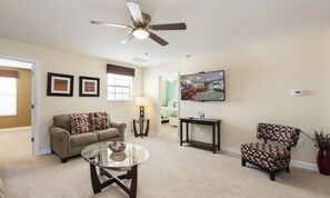 A living room with a sofa, a coffee table, a patterned armchair, a TV, and a ceiling fan. - 8BR Championsgate Cinema, Arcade & Private Pool (Davenport)