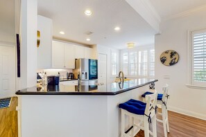 A modern kitchen with white cabinetry, a central island with bar stools, and a window with blinds. - Elevated Condo With Wraparound Porch at Reunion (Kissimmee)