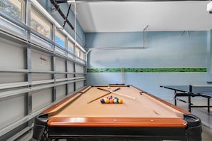 Ein Billardtisch mit Kugeln und Spielstöcken, ein Tischtennis-Tisch und ein Fenster mit Jalousien.